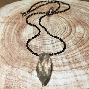 May include: A pendant necklace featuring a faceted, teardrop-shaped pendant. The pendant is a light brown colour and hangs from a delicate black beaded chain. The necklace is fastened with a silver clasp. The background is a wooden surface.