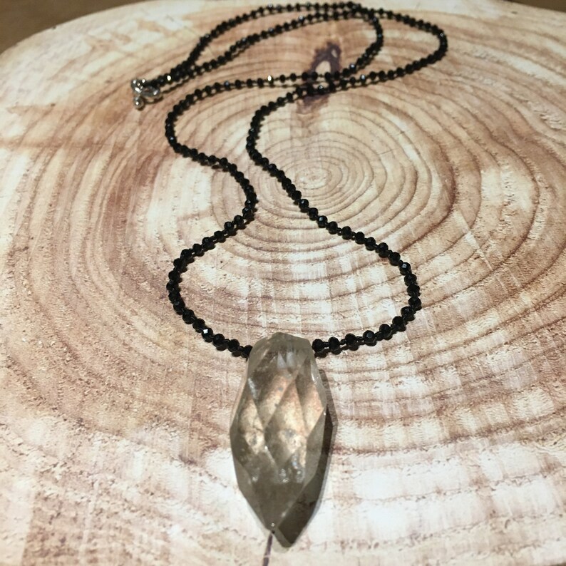 May include: A pendant necklace featuring a faceted, teardrop-shaped pendant. The pendant is a light brown colour and hangs from a delicate black beaded chain. The necklace is fastened with a silver clasp. The background is a wooden surface.