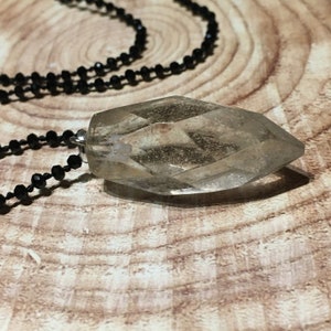 May include: A clear, faceted crystal pendant necklace with a black beaded chain. The crystal is a light, translucent colour and is shaped like a long, pointed gem. The necklace is displayed on a wooden surface.