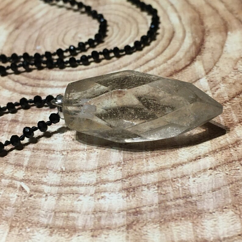 May include: A clear, faceted crystal pendant necklace with a black beaded chain. The crystal is a light, translucent colour and is shaped like a long, pointed gem. The necklace is displayed on a wooden surface.