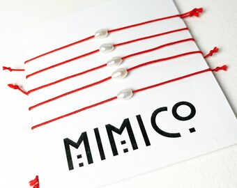 May include: Six red string bracelets with white pearl charms. The bracelets are tied together and the brand name "MIMICO" is printed on a white card.