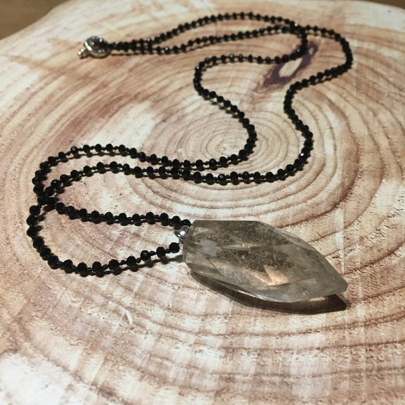 May include: A black beaded necklace featuring a large, clear, faceted crystal pendant. The pendant is a light brown colour and has a unique, elongated shape. The necklace is displayed on a wooden surface.