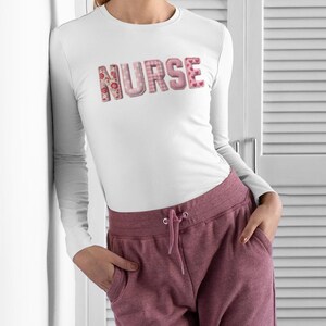 Peut inclure: T-shirt blanc à manches longues avec le mot "NURSE" en rose, motifs floraux et vichy. La personne porte un pantalon de survêtement bordeaux. Le t-shirt est de style col rond.