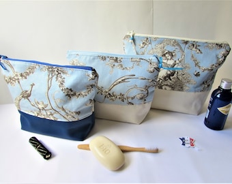 May include: Three toiletry bags in varying sizes. The bags feature a light blue fabric with a brown and white pastoral design. The bottom of the bags are a solid navy blue or cream color. A tube of lipstick, a bar of soap, and a toothbrush are also visible.