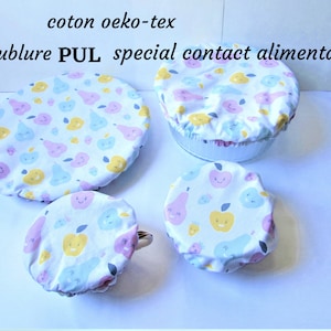 May include: Four reusable fabric bowl covers in varying sizes, featuring a white background with a repeating pattern of smiling yellow apples, pink pears, and light blue fruits. The text reads: coton oeko-tex, doublure PUL, special contact alimentaire.