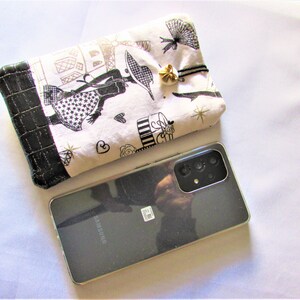 May include: A black Samsung smartphone next to a white and black patterned pouch. The pouch features a black border and a gold-colored charm. The pouch has a design of a woman, Eiffel Tower, and other Parisian elements.