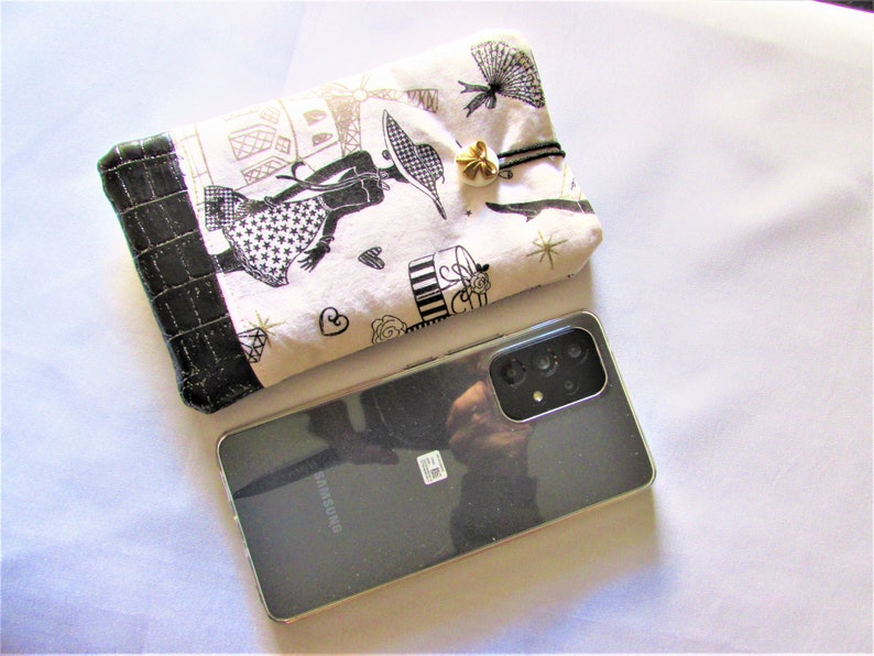 May include: A black Samsung smartphone next to a white and black patterned pouch. The pouch features a black border and a gold-colored charm. The pouch has a design of a woman, Eiffel Tower, and other Parisian elements.
