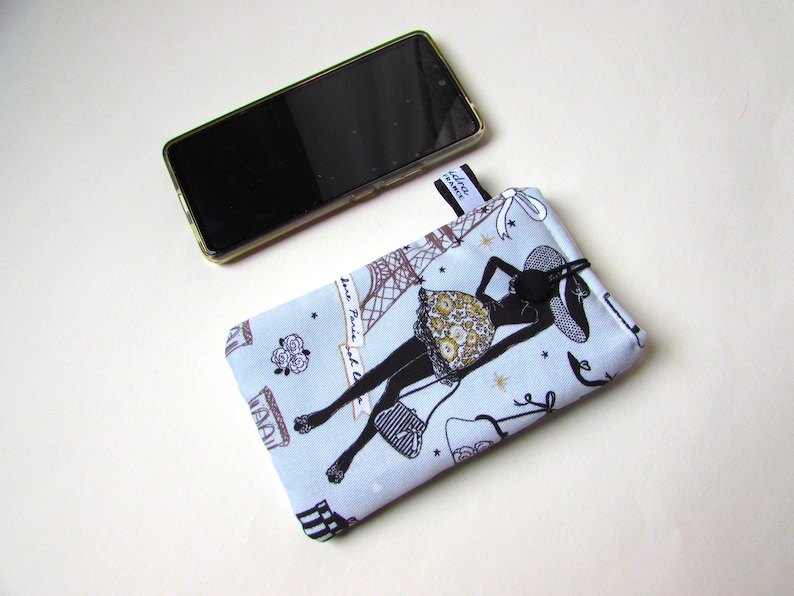 May include: A light blue phone case with a black silhouette of a woman in a hat, Eiffel Tower, and the text "Love Paris". A black smartphone rests above the case. The case has a black tag that says "Idra France".