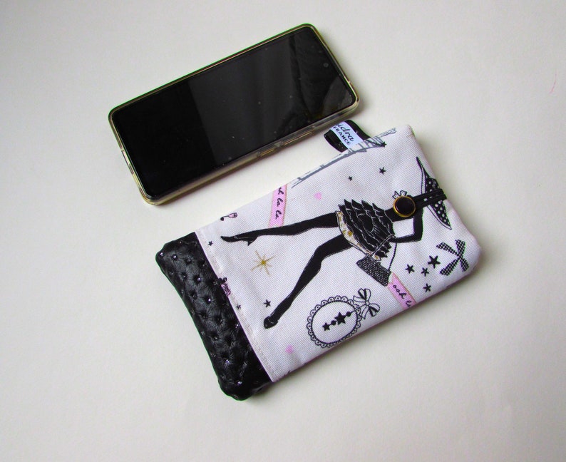 May include: A white and black fabric phone case with a black leather bottom. The case features a black silhouette of a woman in a dress, with pink accents and text. A black smartphone is next to the case.