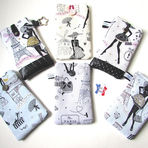 May include: A collection of fabric phone cases with Parisian-themed designs. The cases feature illustrations of the Eiffel Tower, fashion figures, and other Parisian motifs in black and white, with accents of gold and black. Each case has a black leather-like bottom.