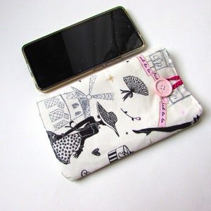 May include: A cream-colored phone case with a black and white Parisian-themed print featuring a woman, windmill, and other illustrations. The case has a pink button closure with a pink and red cord. A black smartphone is placed on top.