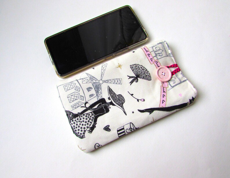 May include: A cream-colored phone case with a black and white Parisian-themed print featuring a woman, windmill, and other illustrations. The case has a pink button closure with a pink and red cord. A black smartphone is placed on top.