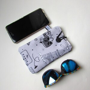 May include: A light gray glasses case with a black and white Parisian design, a black button closure, and a pair of blue mirrored sunglasses. A smartphone rests above the case.