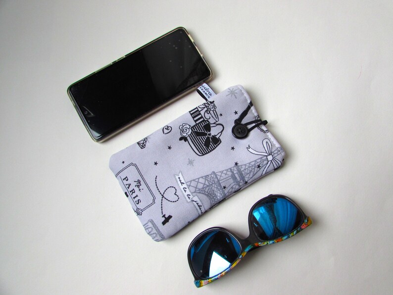 May include: A light gray glasses case with a black and white Parisian design, a black button closure, and a pair of blue mirrored sunglasses. A smartphone rests above the case.