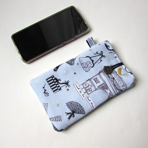 May include: A light blue fabric phone case with a black and white Parisian theme print, featuring the Eiffel Tower and other Parisian motifs. The case has a black elastic closure and a small tag. A smartphone rests beside the case.