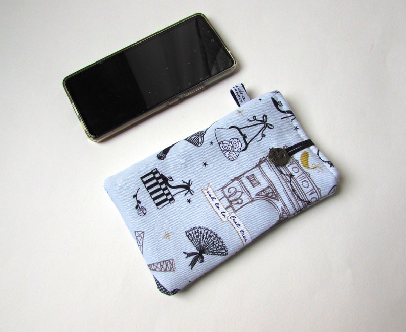 May include: A light blue fabric phone case with a black and white Parisian theme print, featuring the Eiffel Tower and other Parisian motifs. The case has a black elastic closure and a small tag. A smartphone rests beside the case.