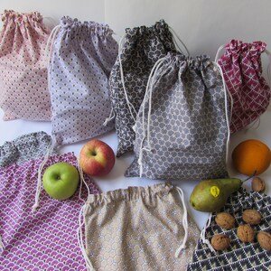 Pode incluir: Uma coleção de sacos reutilizáveis com cordão em várias cores e padrões. Os sacos são feitos de tecido e apresentam um fecho de cordão branco. Frutas e nozes são dispostas ao redor dos sacos, incluindo maçãs, uma laranja, uma pêra e nozes.