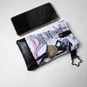 May include: A white and black phone case with a black leather bottom. The case features a design of the Eiffel Tower and a woman in a dress. A black smartphone rests next to the case.
