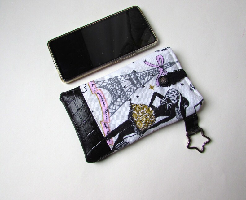 May include: A white and black phone case with a black leather bottom. The case features a design of the Eiffel Tower and a woman in a dress. A black smartphone rests next to the case.
