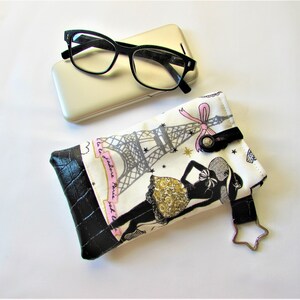 May include: Eyeglasses with black frames rest on a gold phone case, next to a fabric glasses case. The case features a black and white print of the Eiffel Tower and a woman in a dress, with a black leather bottom.