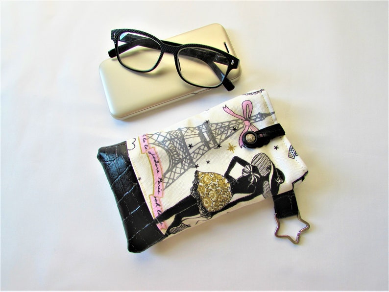 May include: Eyeglasses with black frames rest on a gold phone case, next to a fabric glasses case. The case features a black and white print of the Eiffel Tower and a woman in a dress, with a black leather bottom.