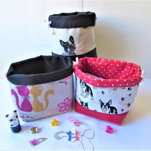 Peut inclure: Trois paniers de rangement en tissu avec des extérieurs noirs, rouges et à motifs. Les paniers présentent des motifs de chats, de chiens et de fleurs. Des accessoires comme des pinces à cheveux et une figurine de panda sont éparpillés.
