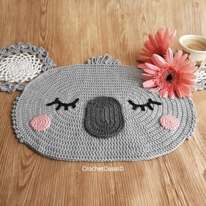 May include: A gray crocheted koala-shaped coaster with pink and black details. The coaster is on a wooden surface with two pink flowers and a coffee cup.