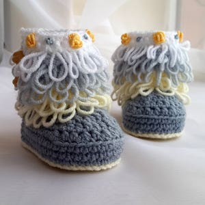 May include: A pair of gray crocheted baby booties with white and yellow crocheted trim. The booties have a ruffled design and are decorated with small yellow buttons.