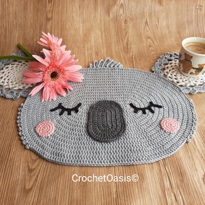 May include: A gray crocheted koala-shaped coaster with pink cheeks and black eyelashes. The coaster is on a wooden surface with a cup of coffee and a pink flower.