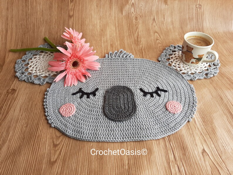 May include: A gray crocheted koala-shaped coaster with pink cheeks and black eyelashes. The coaster is on a wooden surface with a cup of coffee and a pink flower.