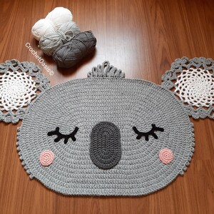 May include: A gray crocheted koala rug with white and gray yarn accents. The rug has a round shape with a face, eyes, and a nose. The rug is on a wooden floor.