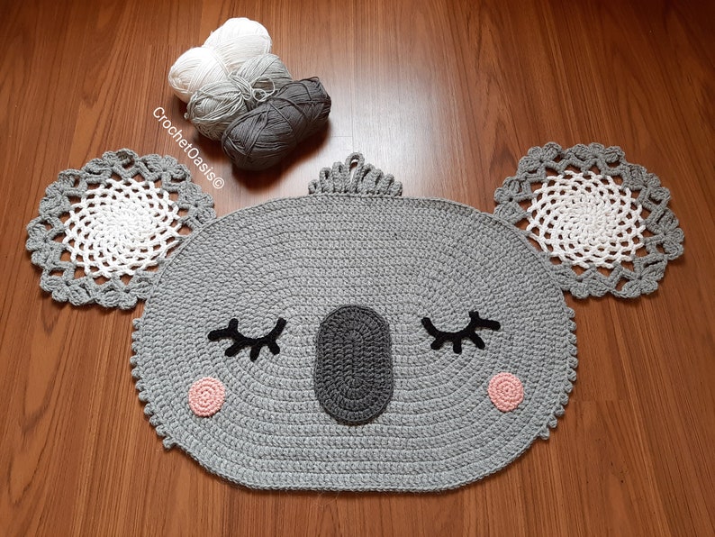 May include: A gray crocheted koala rug with white and gray yarn accents. The rug has a round shape with a face, eyes, and a nose. The rug is on a wooden floor.
