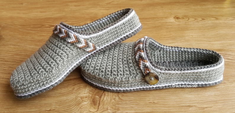 Peut inclure: Une paire de pantoufles en crochet gris avec des bordures blanches et des accents &agrave; motifs brun et blanc. Une pantoufle a un d&eacute;tail bouton.