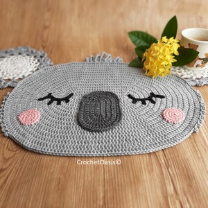 May include: A gray crocheted koala-shaped coaster with black eyes, a black nose, and pink cheeks. The coaster is on a wooden surface with a yellow flower and a cup of coffee.