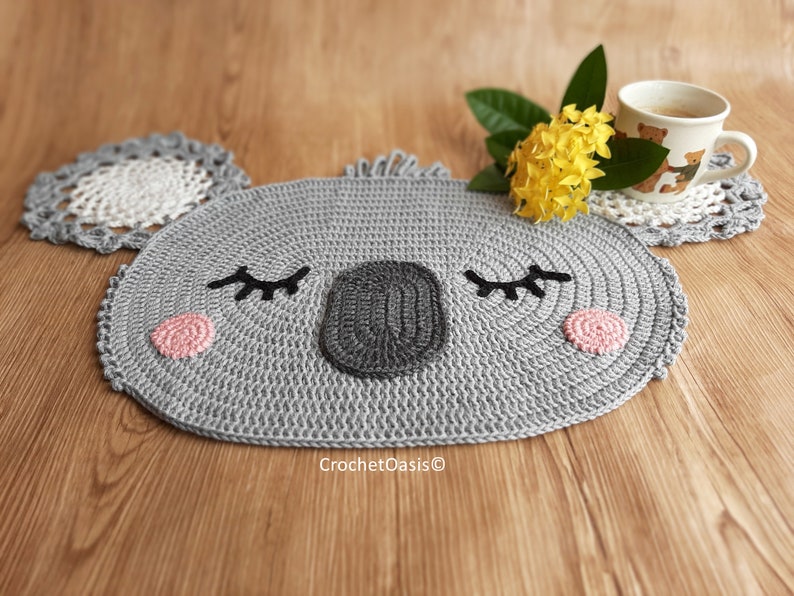 May include: A gray crocheted koala-shaped coaster with black eyes, a black nose, and pink cheeks. The coaster is on a wooden surface with a yellow flower and a cup of coffee.