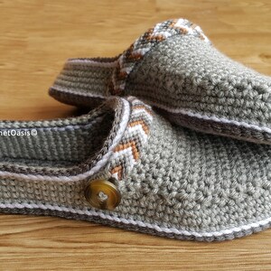 Peut inclure: Une paire de pantoufles en crochet gris avec des bordures blanches et brunes. Les pantoufles ont un bouton sur le dessus et le texte "CrochetOasis&copy;" est visible sur le dessous d'une pantoufle.