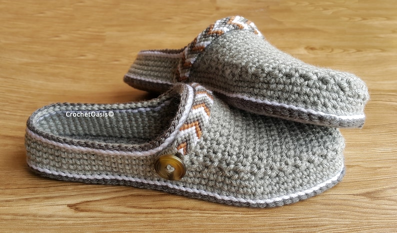 Peut inclure: Une paire de pantoufles en crochet gris avec des bordures blanches et brunes. Les pantoufles ont un bouton sur le dessus et le texte "CrochetOasis&copy;" est visible sur le dessous d'une pantoufle.