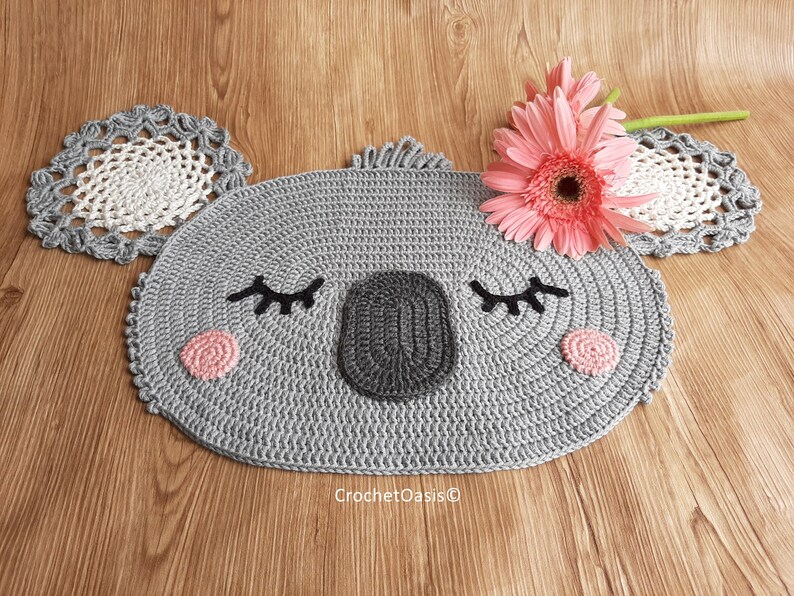 May include: A gray crocheted koala rug with pink cheeks and black eyelashes. The rug is oval shaped and has a white crocheted flower on top.