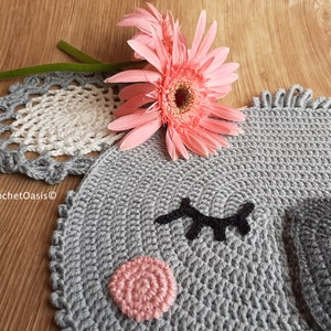 May include: A gray crocheted rug shaped like an elephant's face with black eyelashes, a pink nose, and a gray ear. The rug is on a wooden surface with a pink flower.