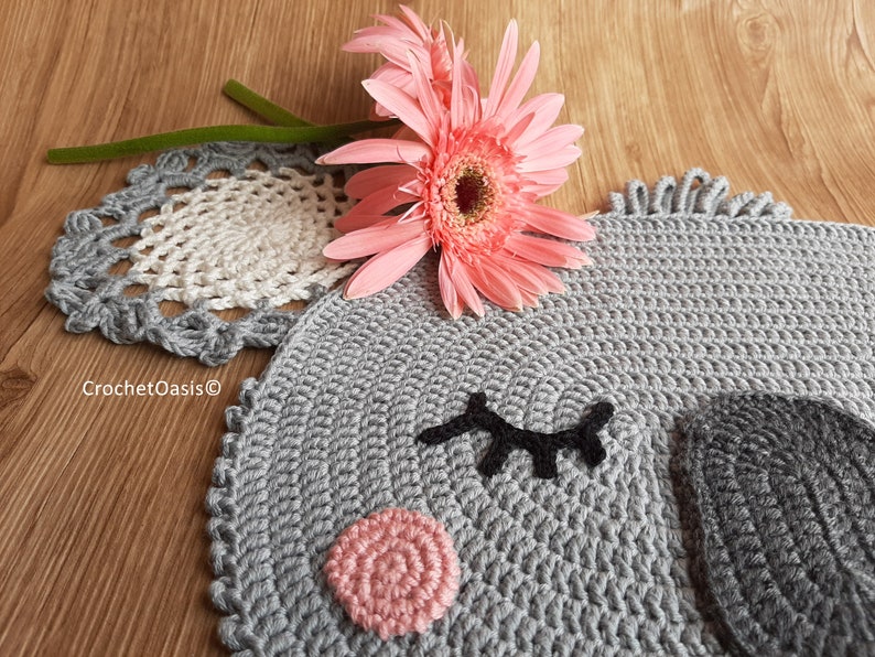 May include: A gray crocheted rug shaped like an elephant's face with black eyelashes, a pink nose, and a gray ear. The rug is on a wooden surface with a pink flower.