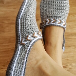 Peut inclure: Paire de pantoufles en crochet gris avec des bordures blanches et brunes. Les pantoufles ont un d&eacute;tail de bouton sur le dessus.