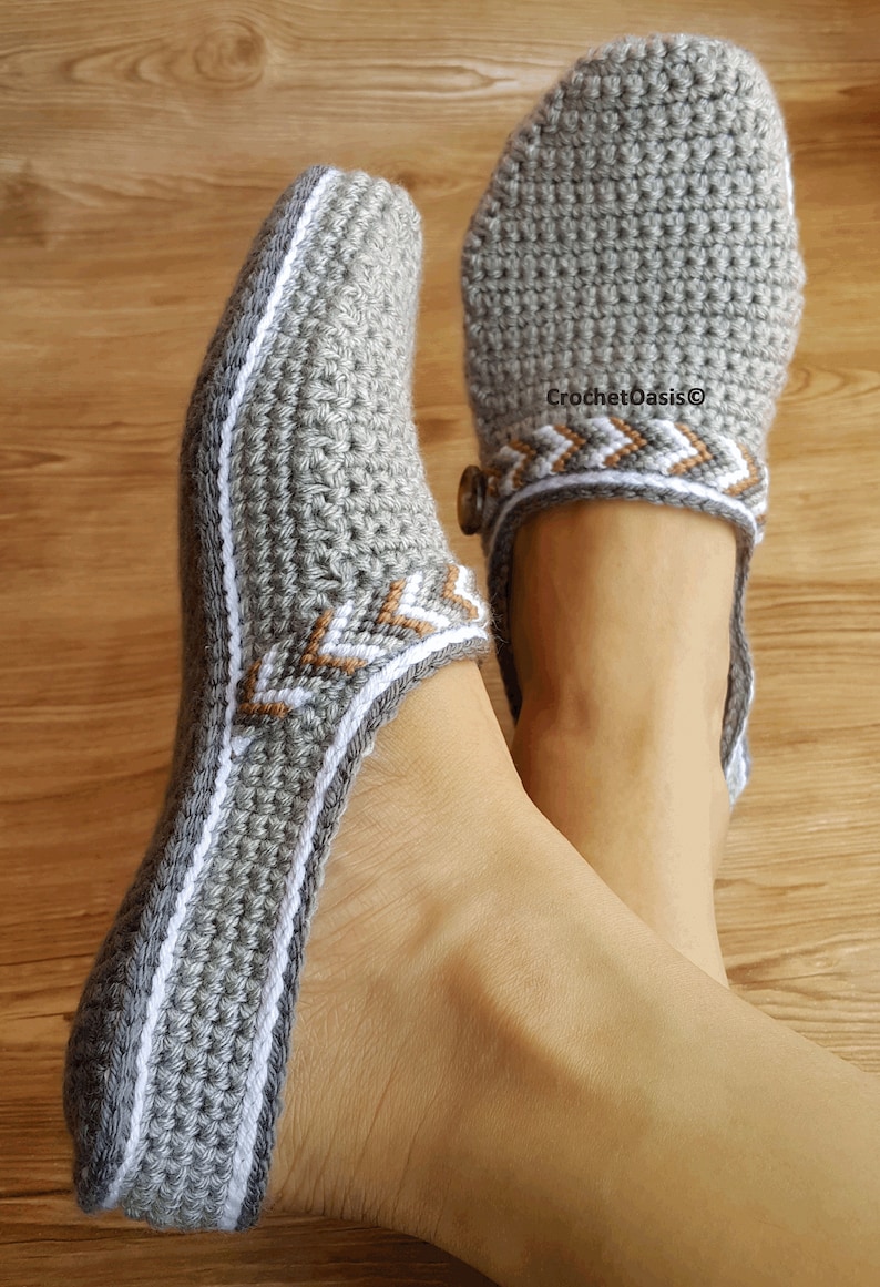 Peut inclure: Paire de pantoufles en crochet gris avec des bordures blanches et brunes. Les pantoufles ont un d&eacute;tail de bouton sur le dessus.
