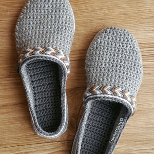 Peut inclure: Une paire de pantoufles en crochet gris avec une bordure blanche et marron. Les pantoufles ont un design en crochet et sont faites avec un fil doux. Le texte "CrochetOasis&reg;" est visible sur la pantoufle de droite.