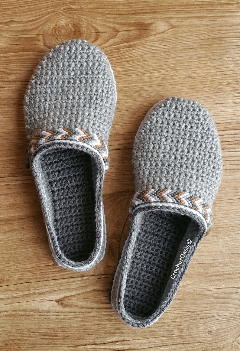 Peut inclure: Une paire de pantoufles en crochet gris avec une bordure blanche et marron. Les pantoufles ont un design en crochet et sont faites avec un fil doux. Le texte "CrochetOasis&reg;" est visible sur la pantoufle de droite.