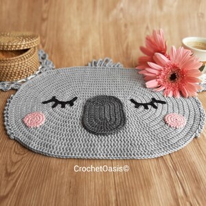 May include: A gray crocheted koala-shaped coaster with pink cheeks and black eyes. The coaster is on a wooden surface with a wicker basket and pink flowers.