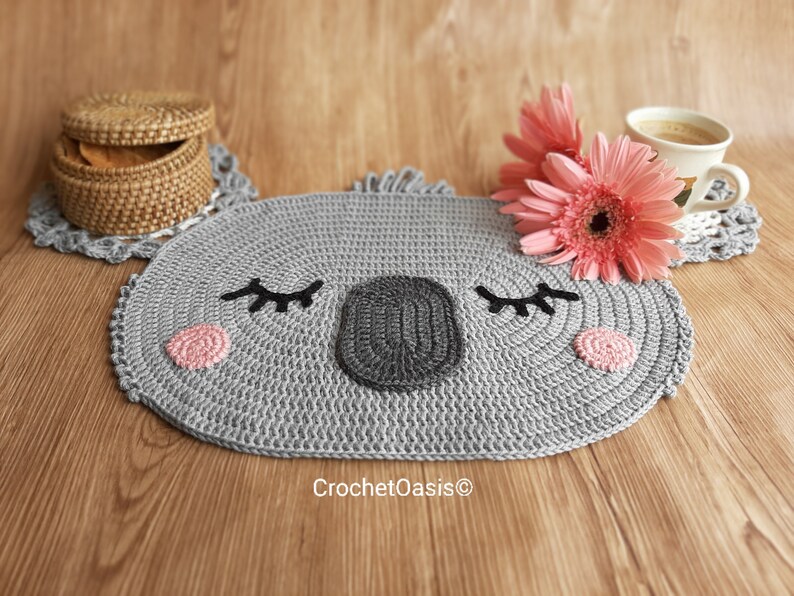 May include: A gray crocheted koala-shaped coaster with pink cheeks and black eyes. The coaster is on a wooden surface with a wicker basket and pink flowers.