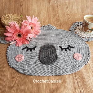 May include: A gray crocheted koala-shaped coaster with pink cheeks and black eyes. The coaster is on a wooden surface with pink flowers and a coffee cup.