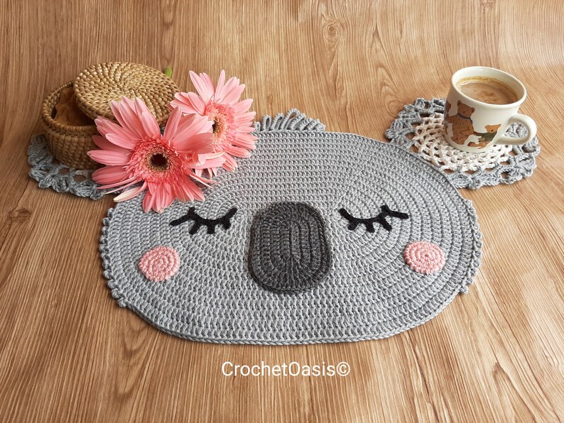 May include: A gray crocheted koala-shaped coaster with pink cheeks and black eyes. The coaster is on a wooden surface with pink flowers and a coffee cup.