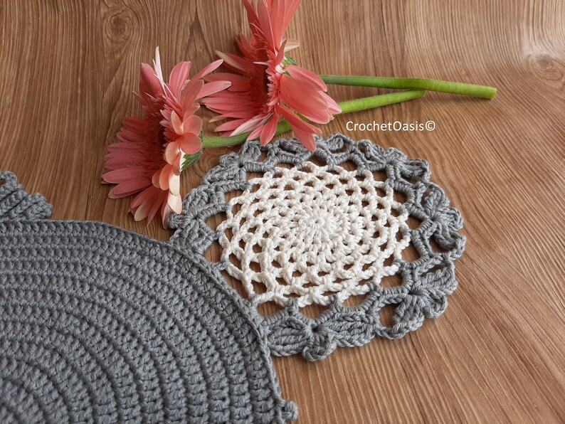 May include: Two crocheted doilies, one gray and one white with a gray border, on a wood surface with pink flowers.