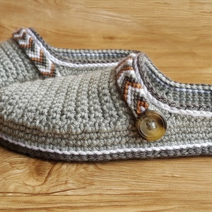 Peut inclure: Une paire de pantoufles en crochet gris avec des bordures blanches et brunes. Les pantoufles ont un bouton sur le dessus et le texte "Crochet Oasis" est visible sur la pantoufle de droite.